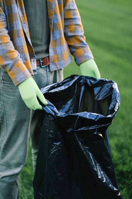 A person standing outdoors on a grassy area, wearing green gloves, a green T-shirt, and a plaid shirt with shades of yellow, blue, and gray. They are holding open a large black rubbish bag made of glo