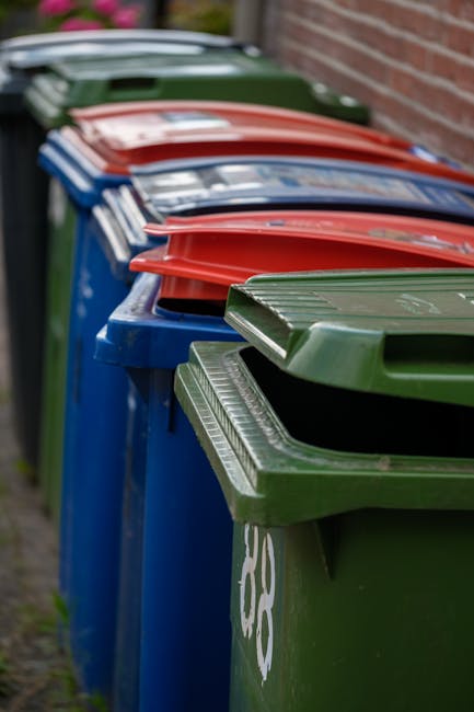 A row of five large wheeled rubbish bins with black plastic bodies and bright yellow lids, aligned against a plain, light gray concrete wall. The bins are positioned on a paved outdoor surface, with t