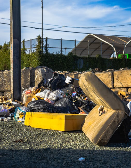 A close-up view of a rubbish accumulation collection on a paved surface outside, featuring a variety of discarded waste including black plastic bin bags, a yellow plastic tray, a worn and stained car 