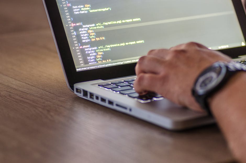 A close-up view of a person's hand typing on a silver laptop keyboard placed on a wooden desk with a natural finish. The person's wrist is adorned with a dark wristwatch with a round face and a textur